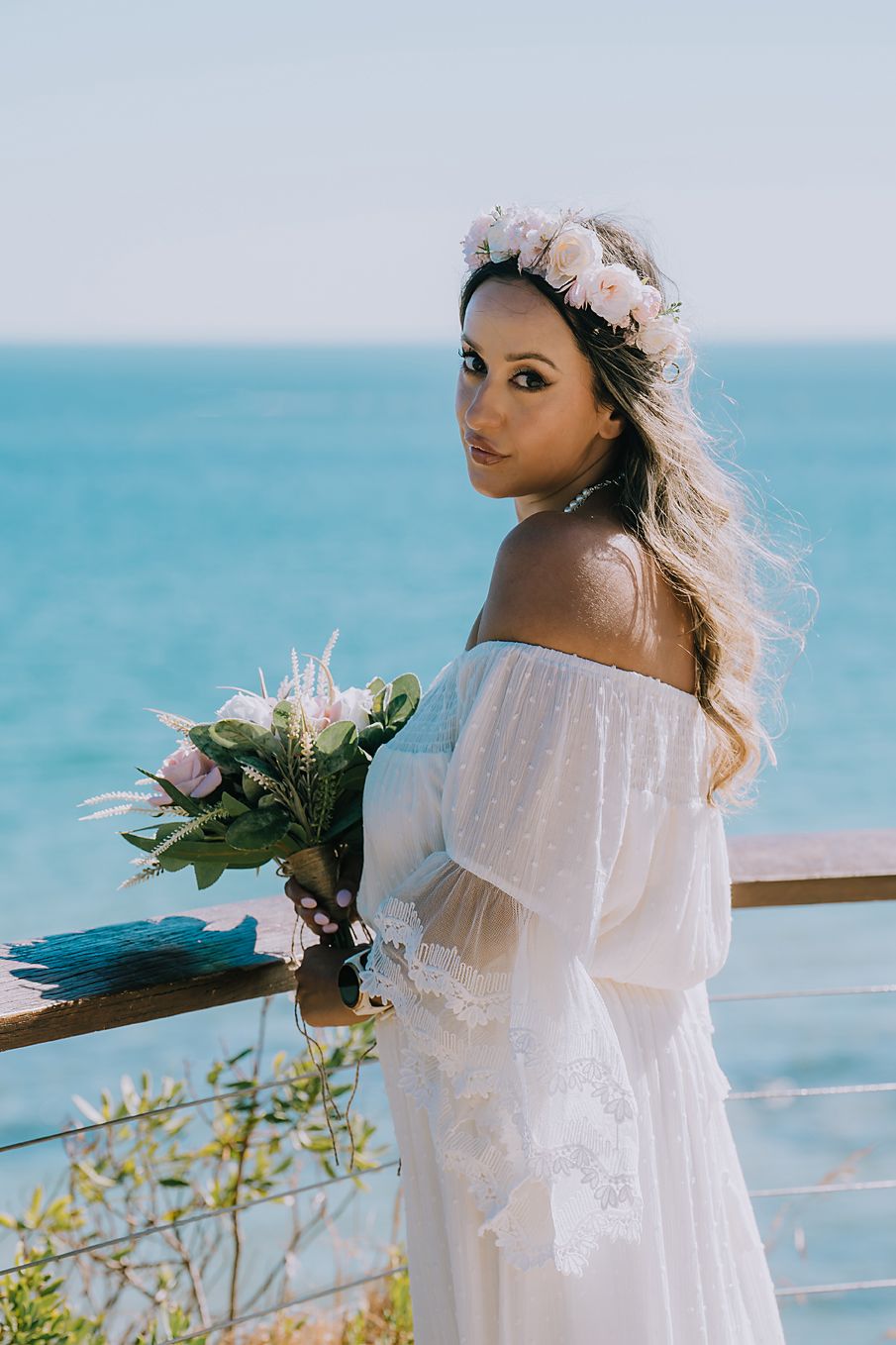 A bride in a flowing white off-shoulder dress poses by the ocean with pink flowers and a floral crown.