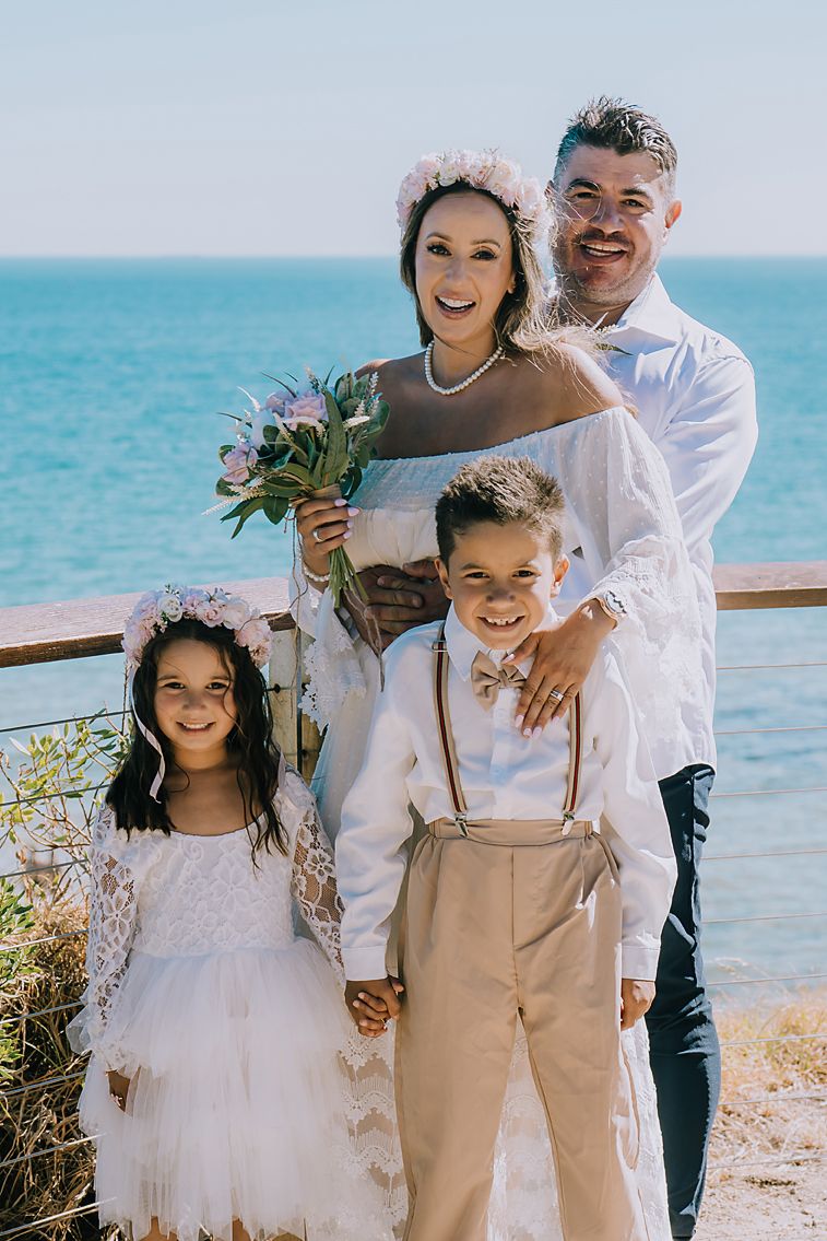 Family wearing white and beige outfits poses together on a beach with turquoise ocean waters in the background.