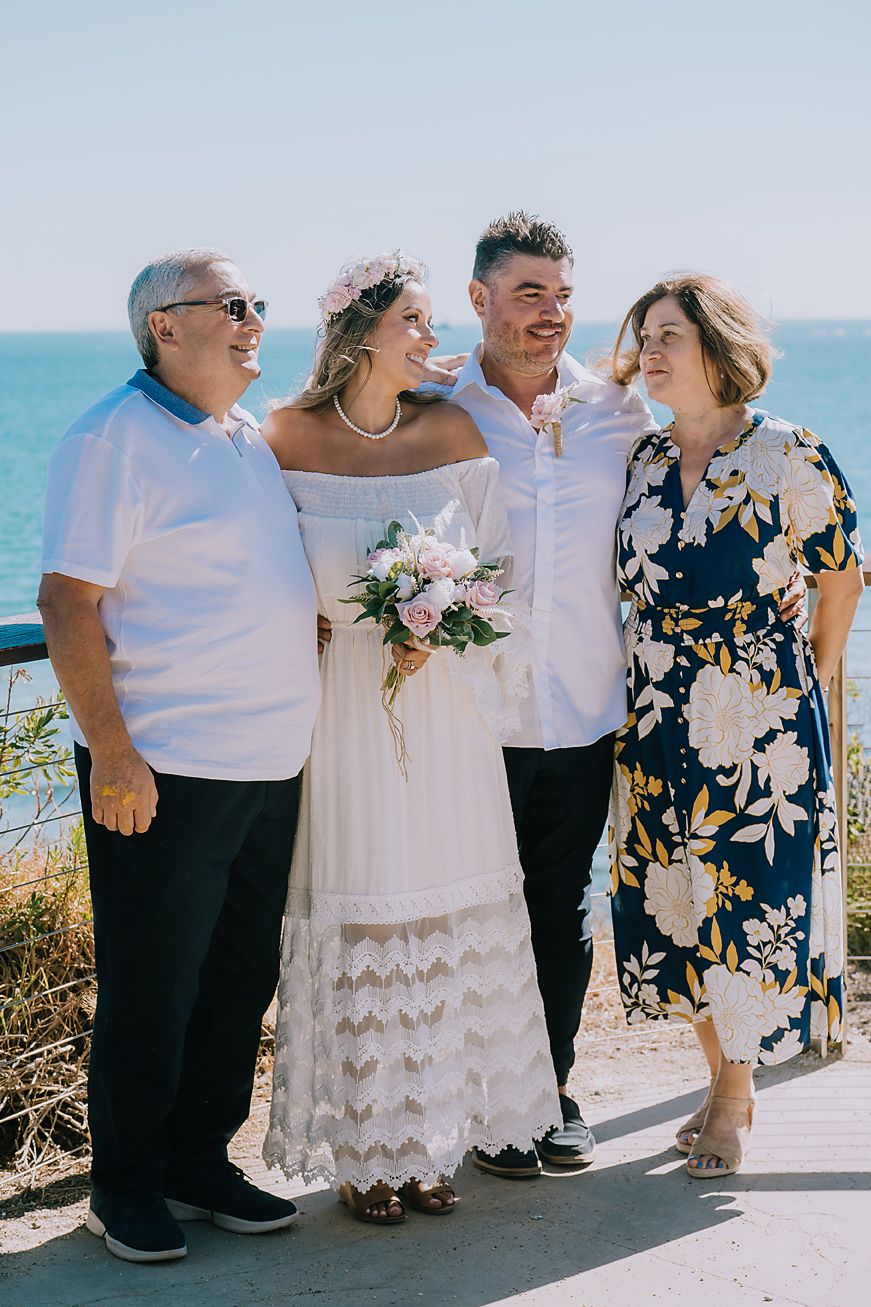 A group enjoying a sunny beachside celebration wearing white and floral outfits against an ocean backdrop.