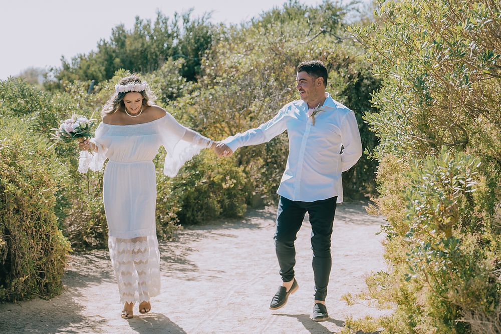 A couple walks hand in hand along a winding sandy path bordered by lush green vegetation on a sunny day.