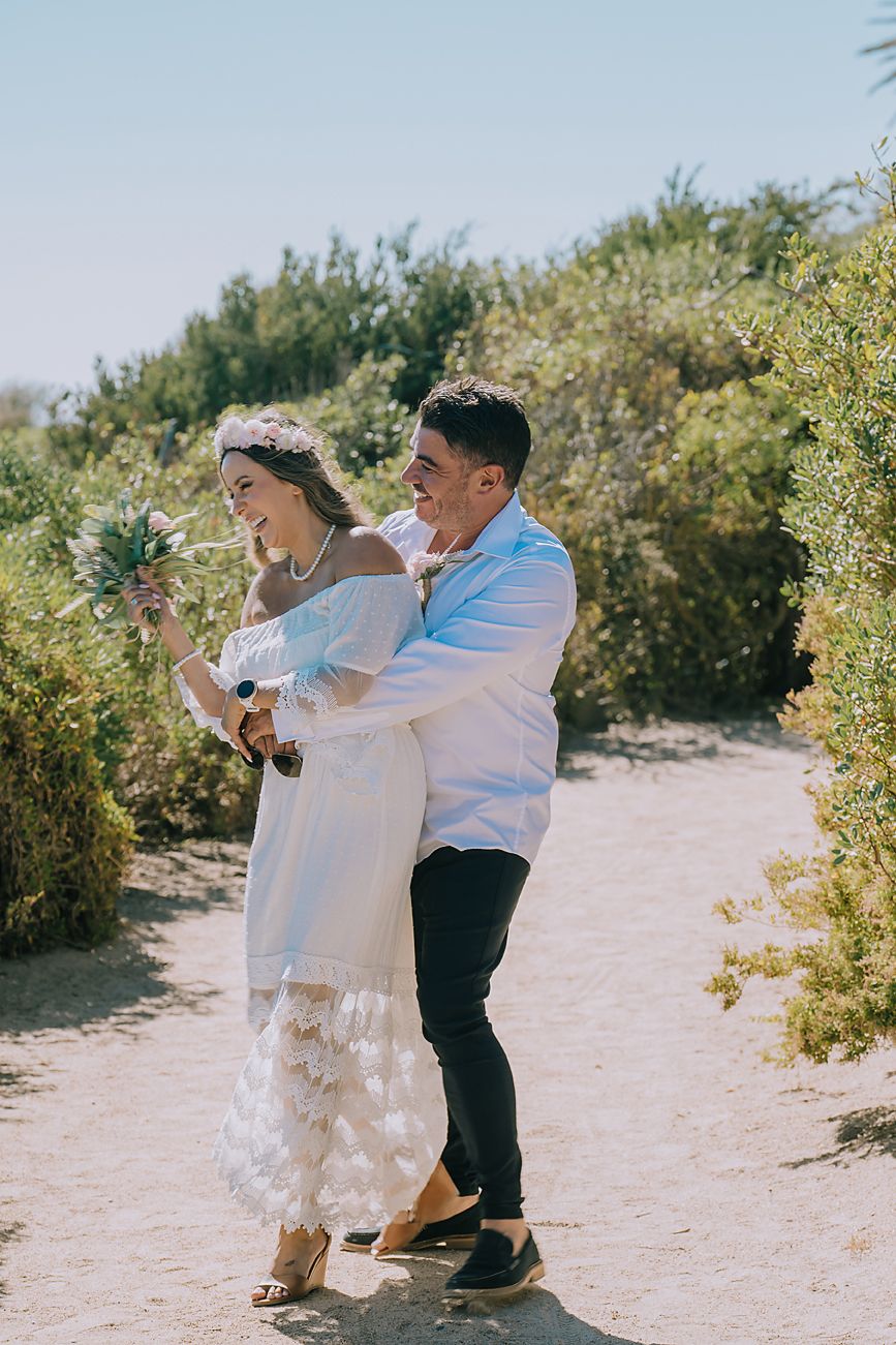 A couple in wedding attire dances and twirls together on a sandy path surrounded by desert brush and greenery.