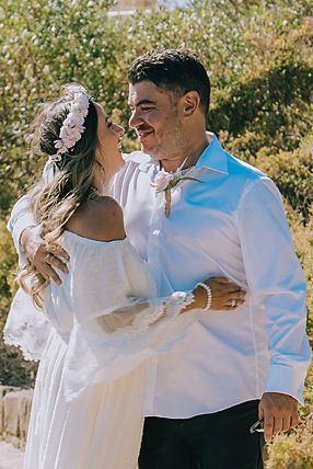 A couple in white attire embraces and dances outdoors on a sunny day with autumn foliage in the background.