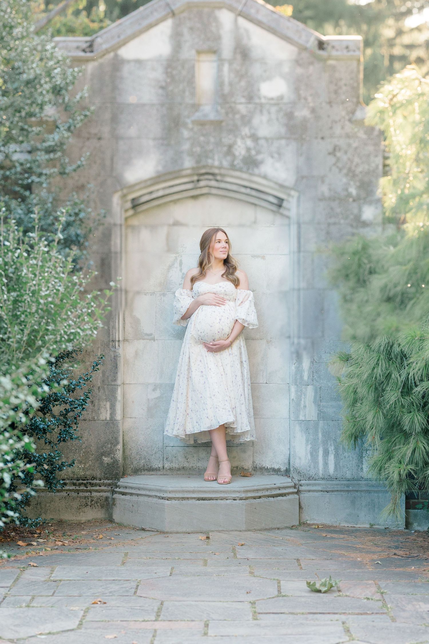 A dreamy maternity photo of an expectant mother in a white dress standing in a stone archway surrounded by greenery.