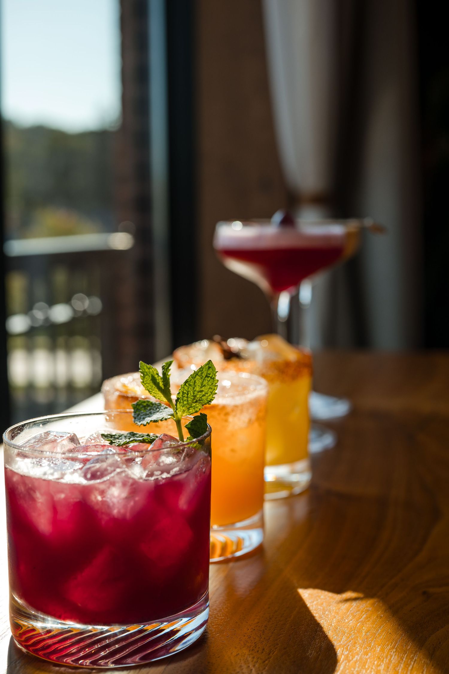 Vibrant craft cocktails with garnishes lined up on a wooden bar in warm lighting, featuring red and orange hues.