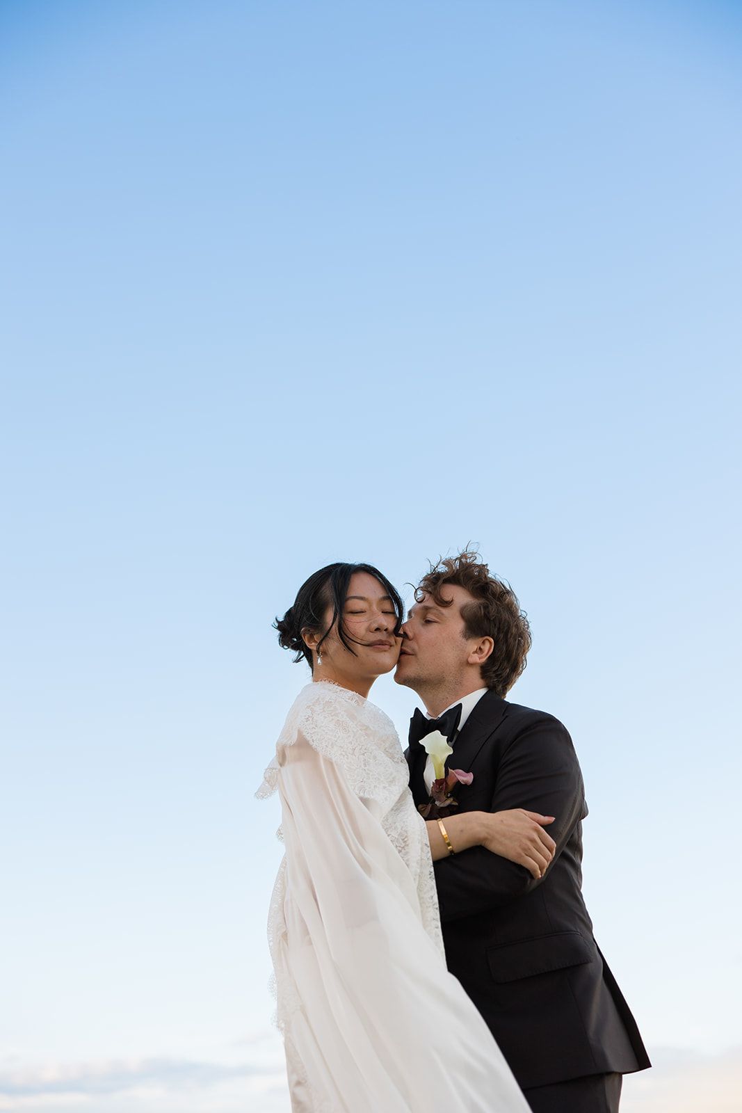A romantic couple shares an intimate kiss against a bright blue sky during their wedding celebration.