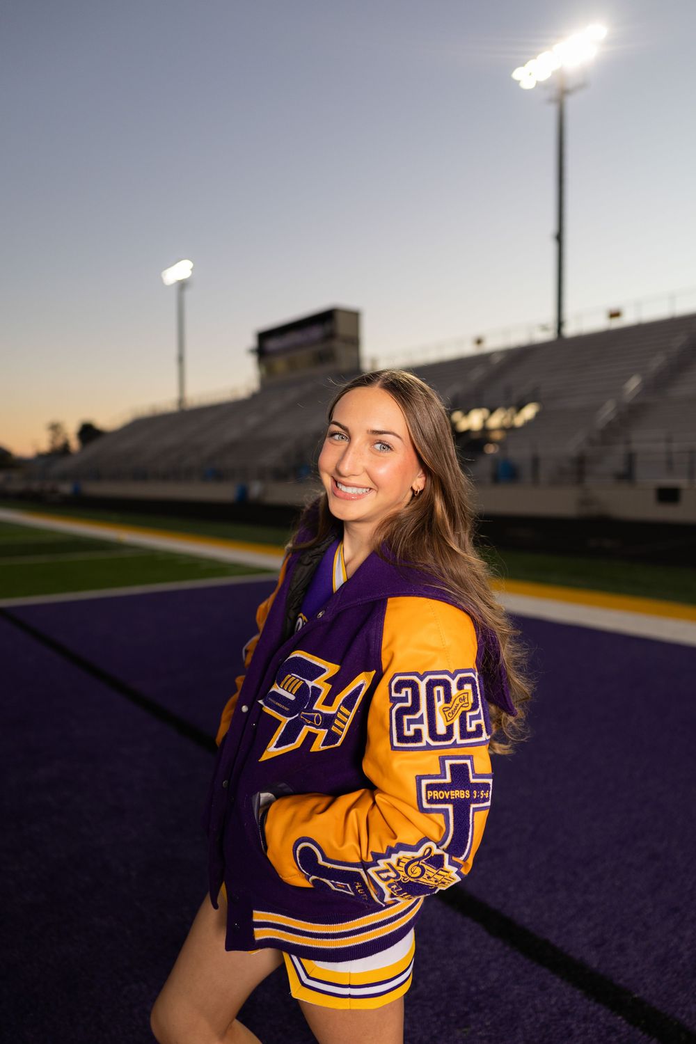 Cheerleader in purple and gold letterman jacket poses on football field at sunset.