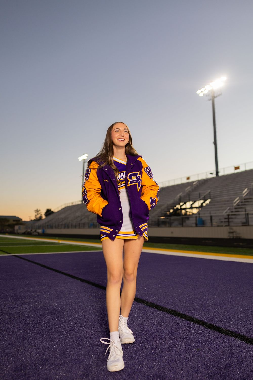 Cheerleader in varsity jacket smiles while standing on purple football field at dusk.