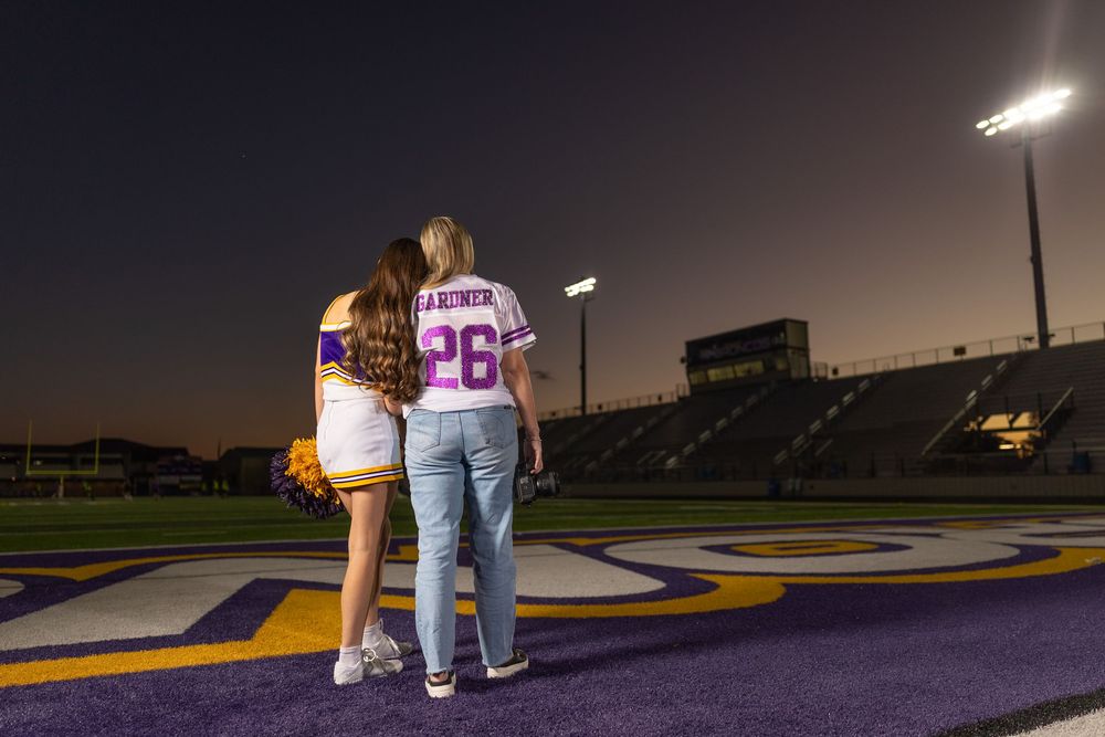 Two people stand together on purple football field at night looking at stadium.