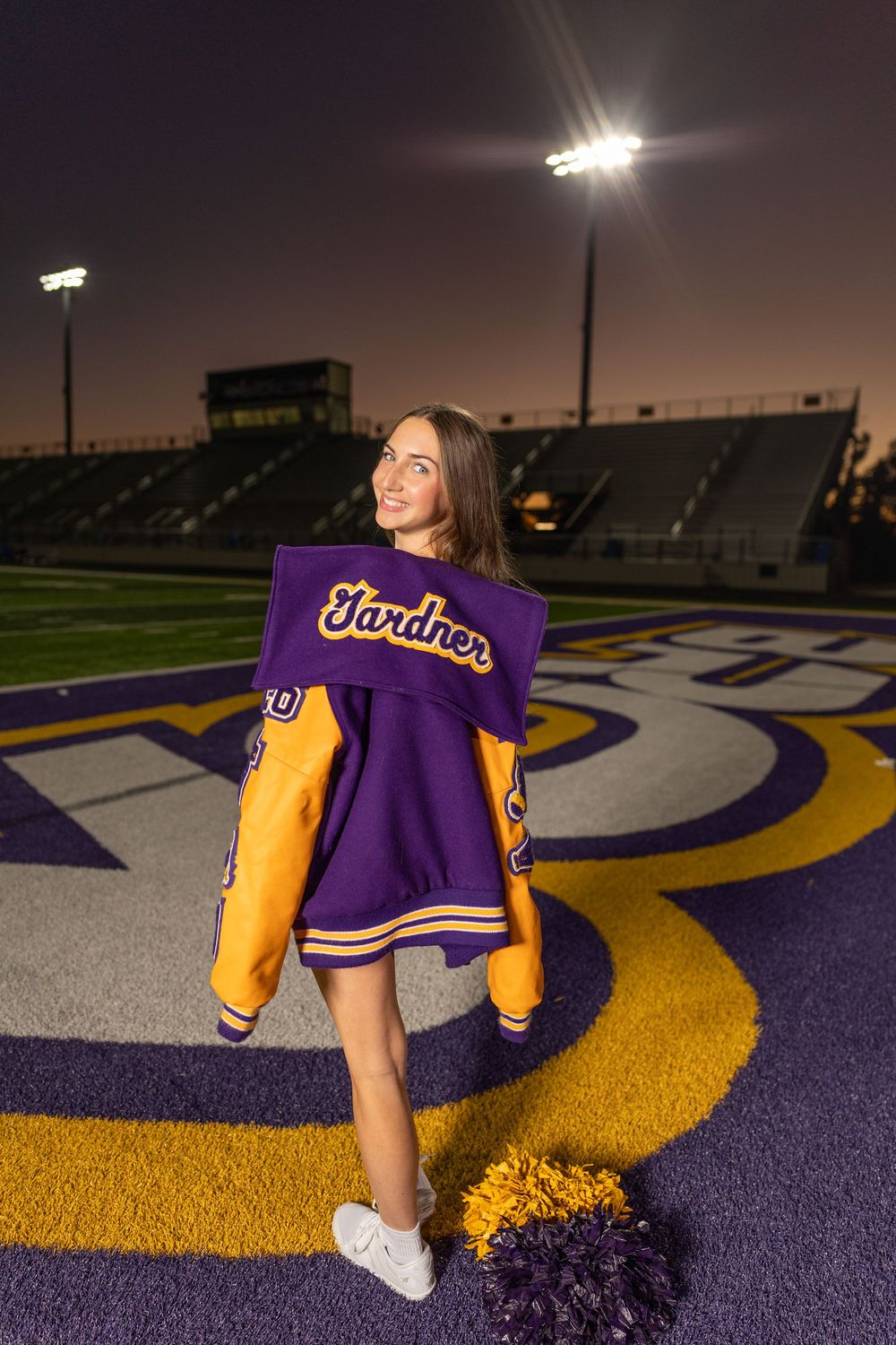 Cheerleader displays purple uniform on illuminated football field at night.