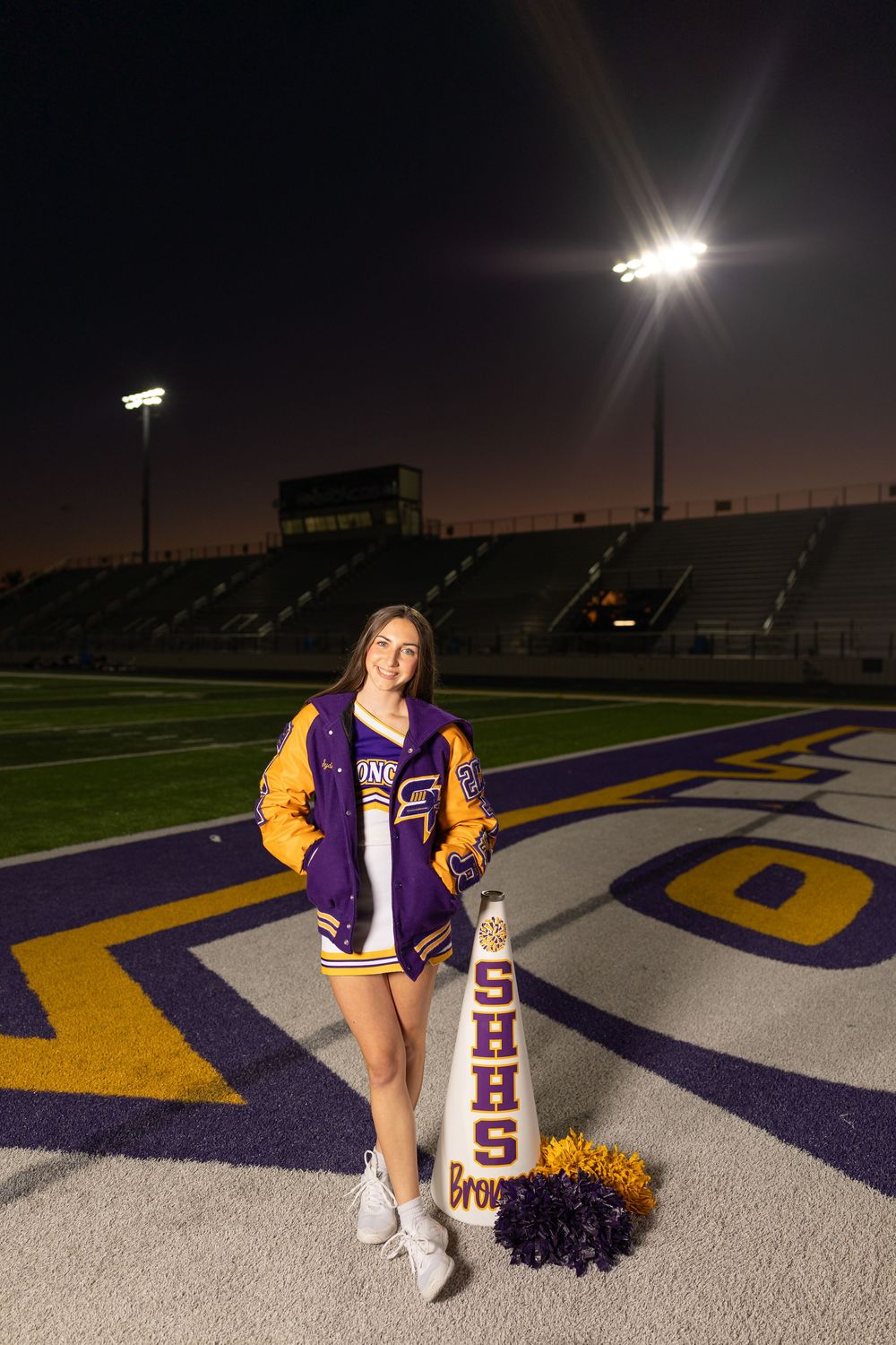Cheerleader in purple and gold varsity jacket poses on football field at night under stadium lights.