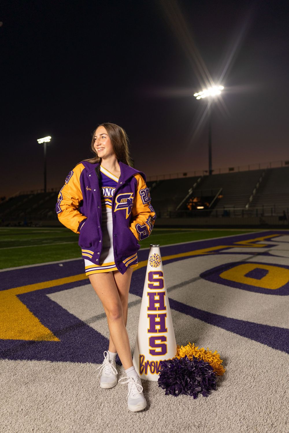 Cheerleader with pom poms sits on stadium field wearing purple and gold varsity jacket.