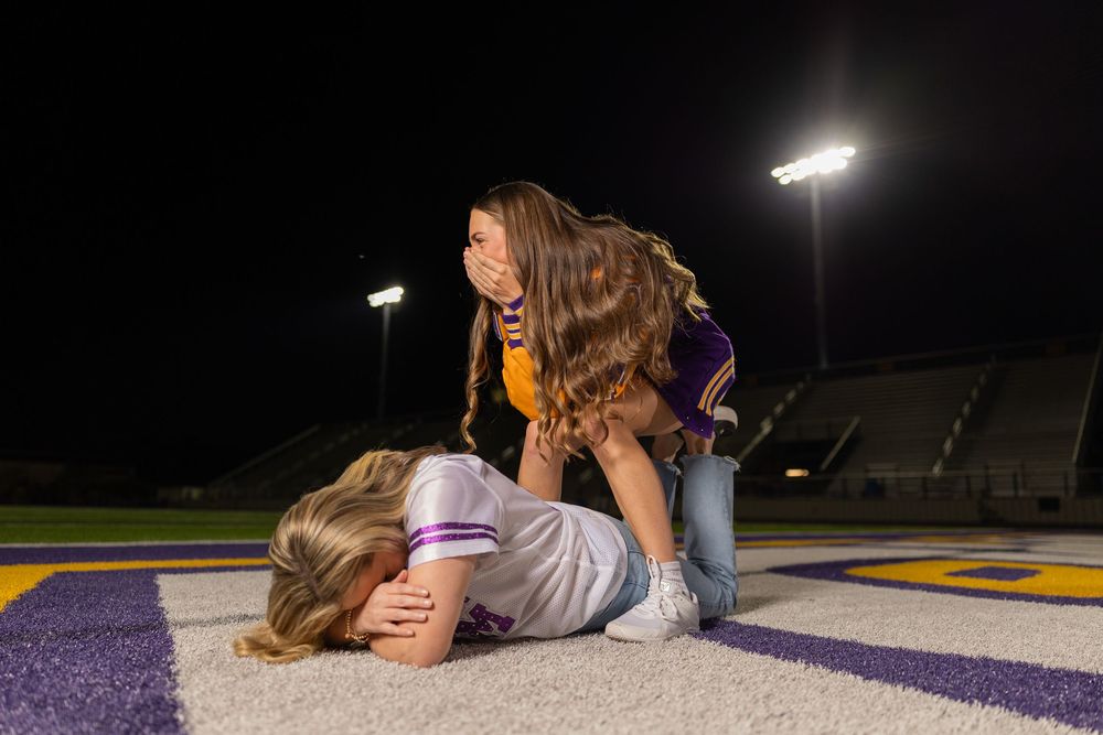 Students in varsity jackets lying on football field turf under stadium lights.