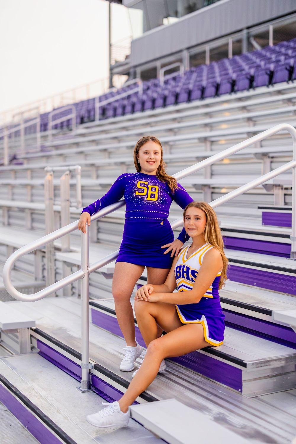 Two cheerleaders in purple uniforms pose on stadium bleachers.
