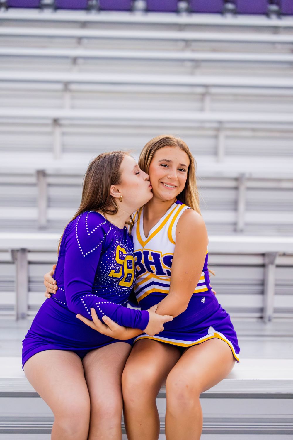 Two cheerleaders in purple uniforms share a friendly moment on stadium bleachers.