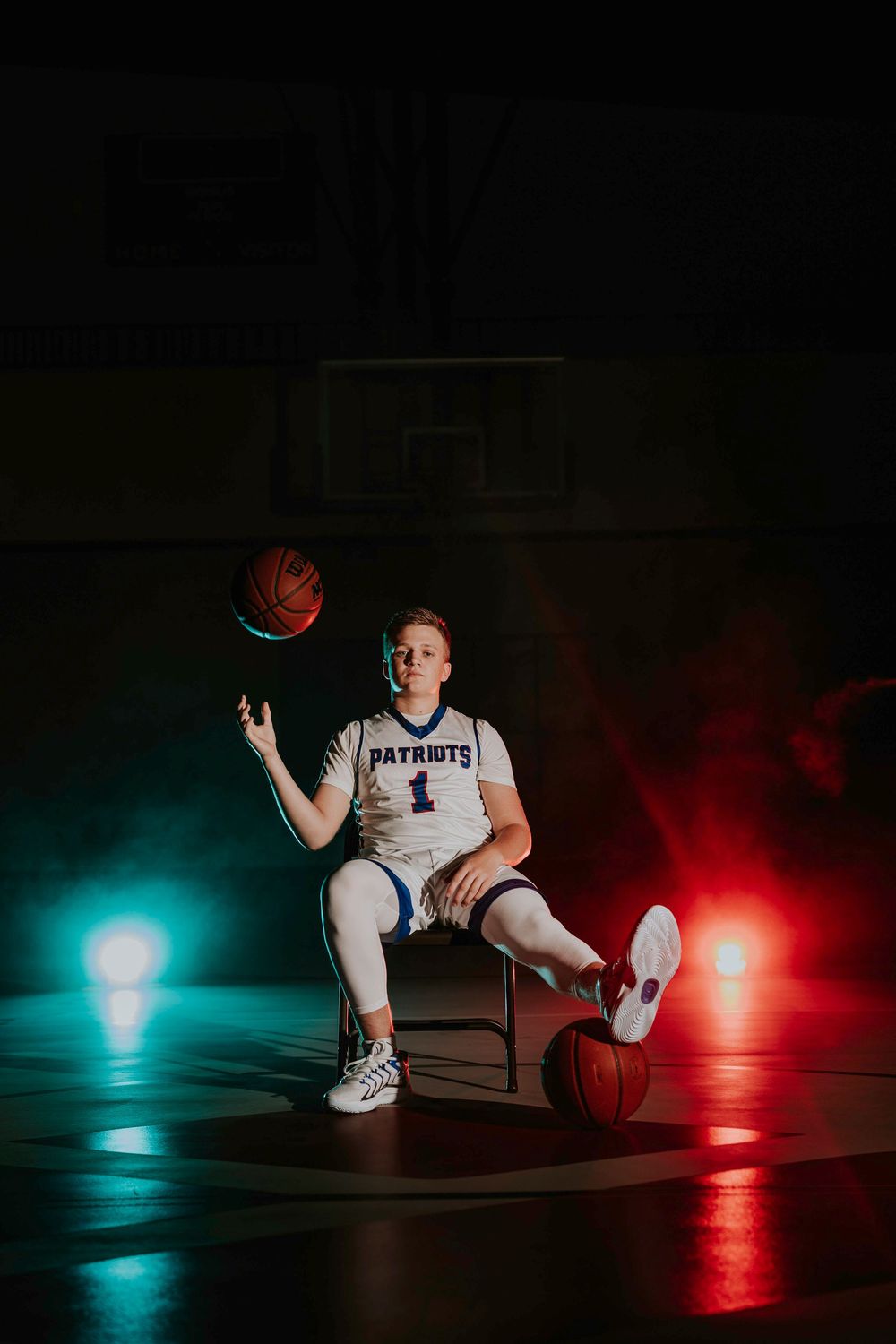 Basketball player in Patriots uniform poses with ball under dramatic red and blue lighting.