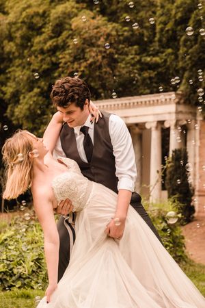 A newlywed couple dances and twirls in a garden setting with floating bubbles around them. Riley dipping Sarah during their photoshoot.