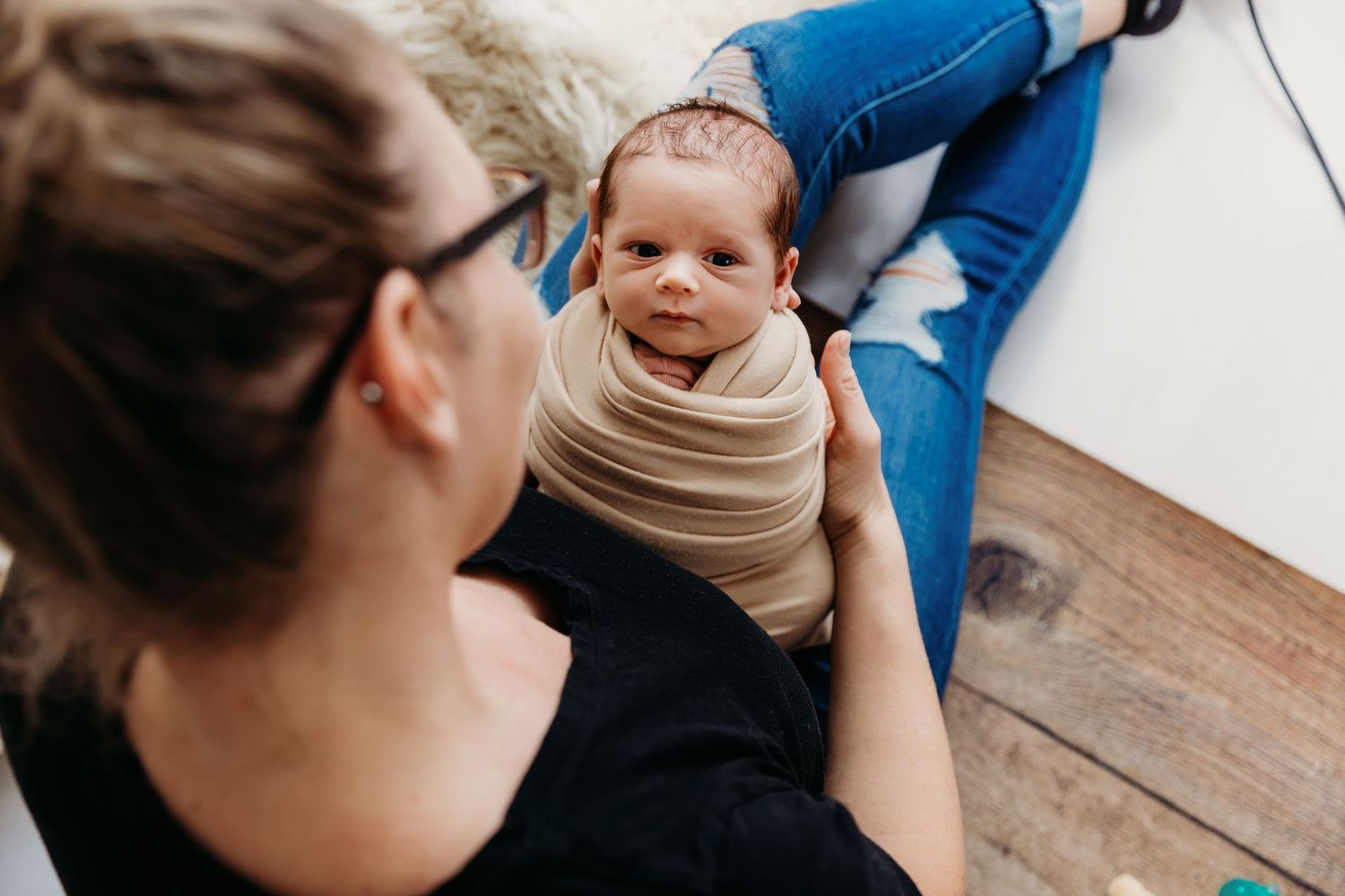 Behind the scenes photographer holding newborn in tan wrap  on wooden floor.
