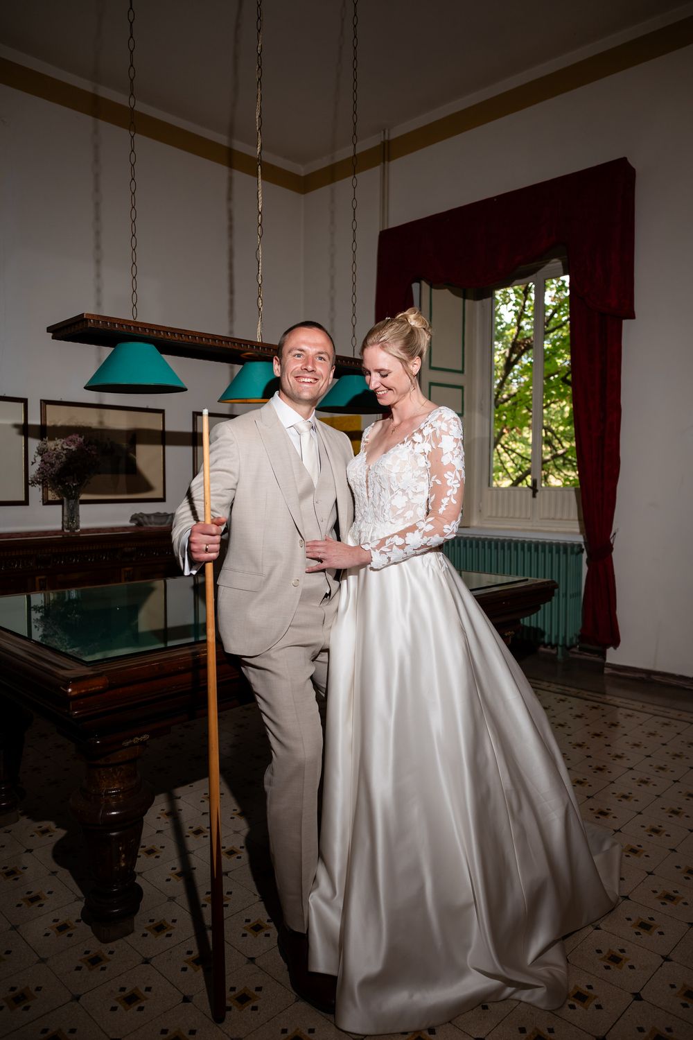 Elegant wedding couple poses in formal attire next to billiards table in classic interior with green lamp overhead.