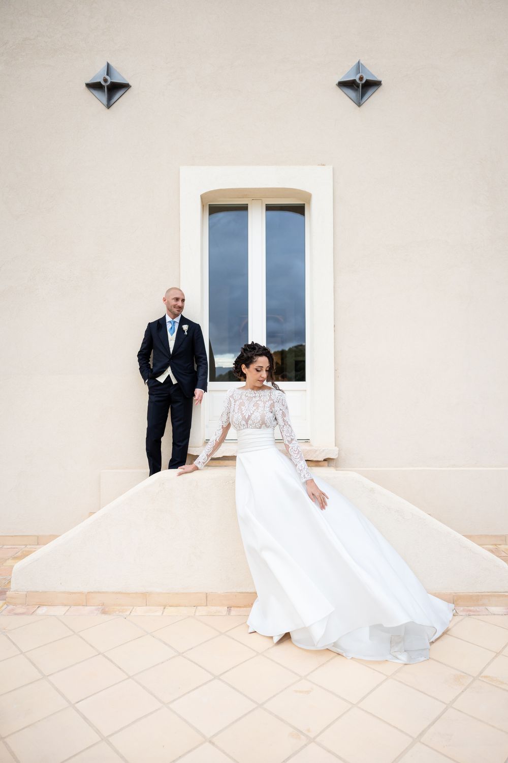 A wedding couple sits playfully on a white tiled terrace against a beige building wall with blue windows.