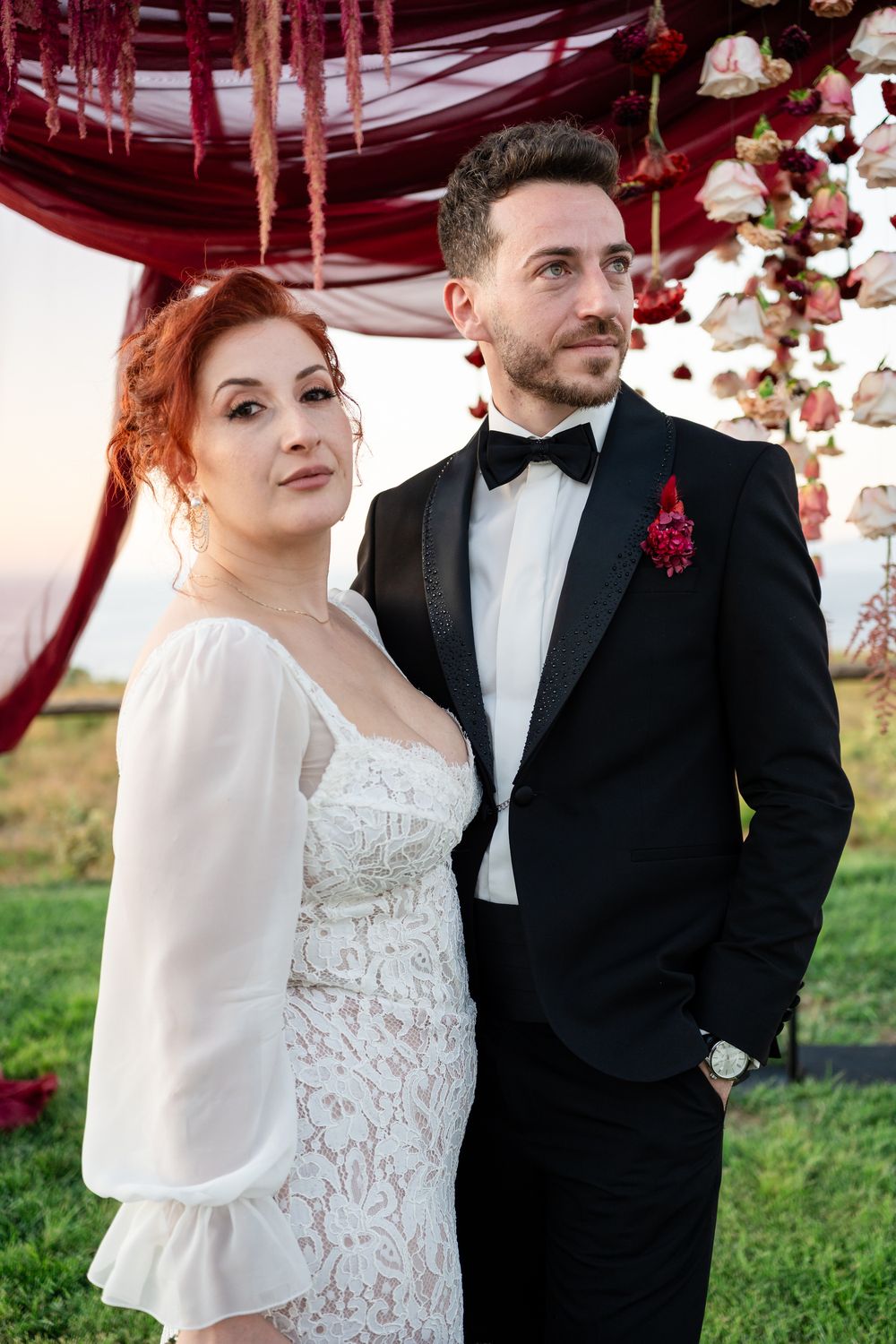 A couple poses outdoors at their wedding under a red draped canopy with floral decorations.