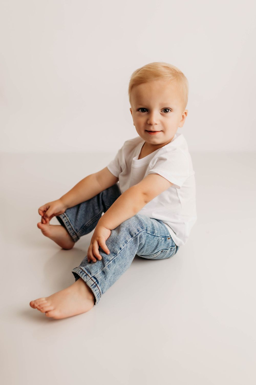 A baby in a white t-shirt and jeans sits and plays on a white studio backdrop in a series of candid poses.