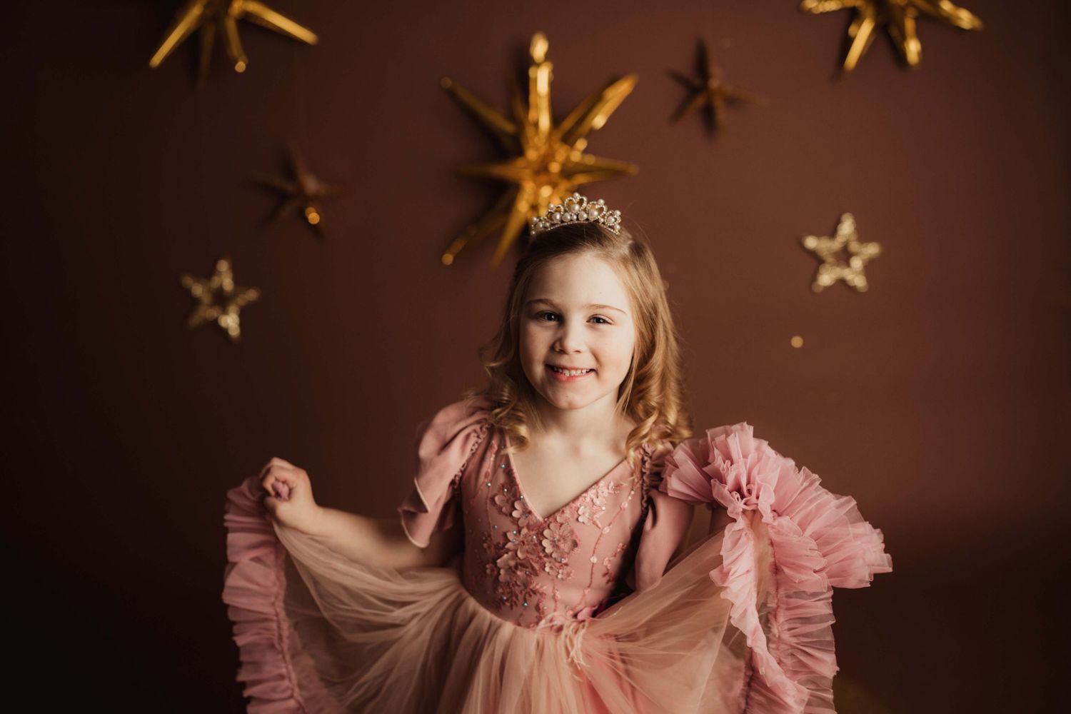 A child wearing a pink princess dress and tiara poses against a dark brown backdrop with gold star decorations.