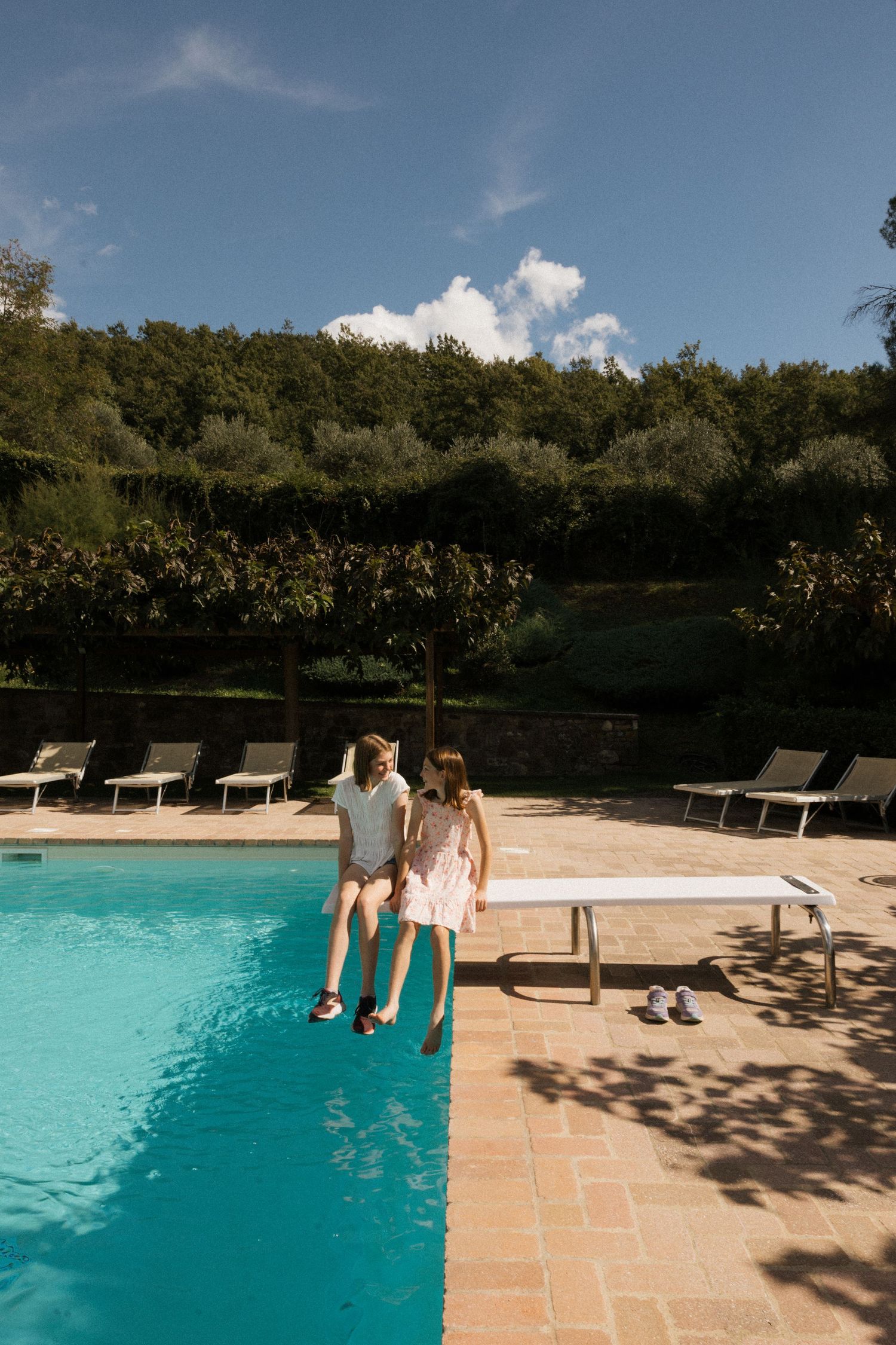 People dangling their feet in a luxurious swimming pool surrounded by lounge chairs on a sunny day.