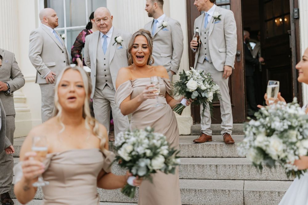 Wedding party members in light gray suits and dresses celebrate joyfully outside an elegant white building.