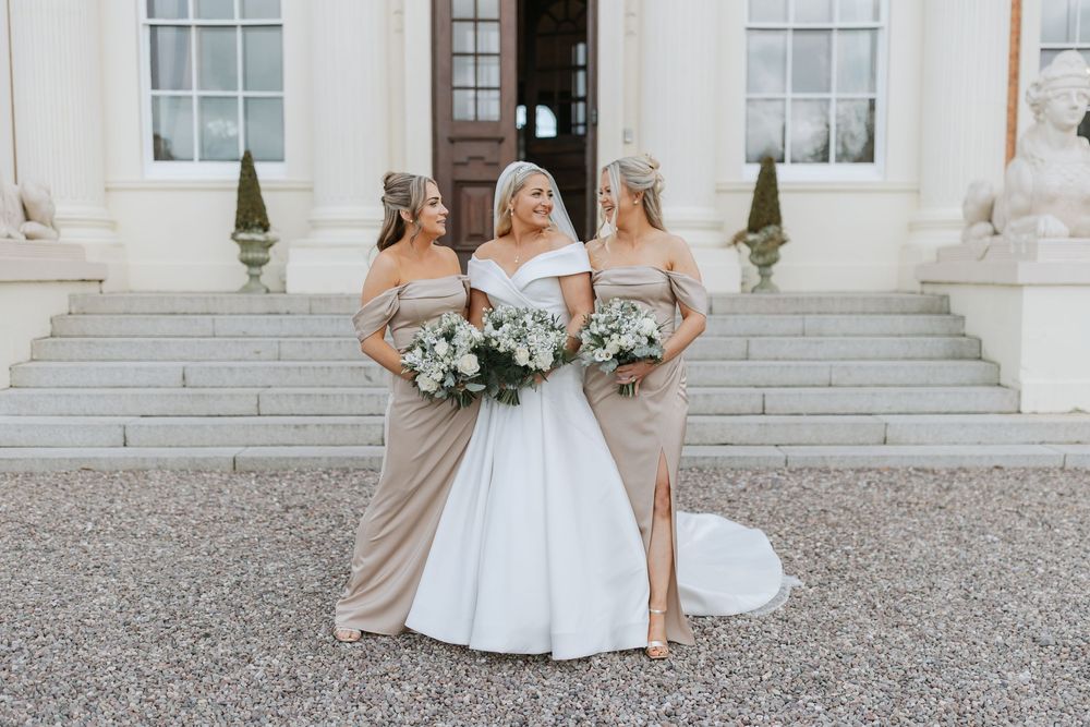 Bridesmaids in neutral-colored dresses pose in front of a grand white mansion with classical architecture.