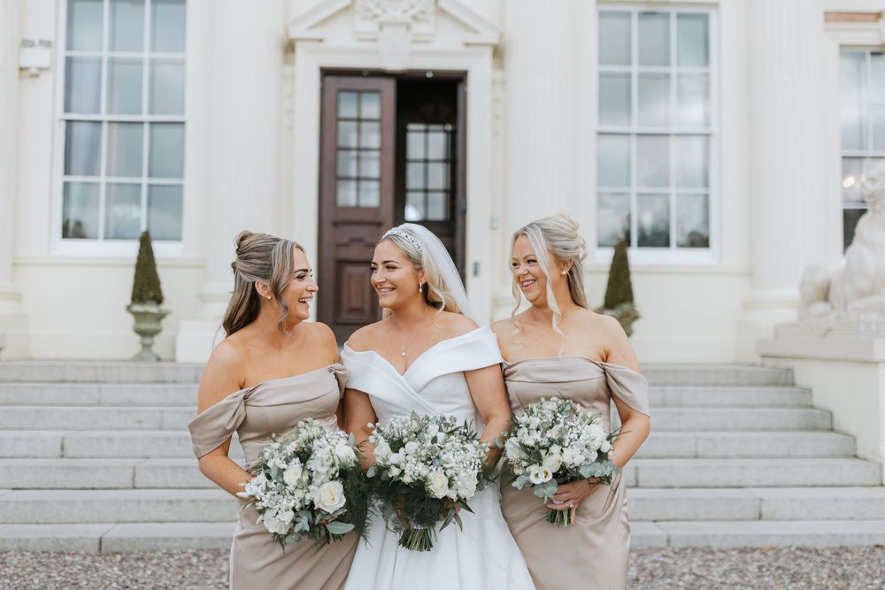 A wedding party in white dresses and suits poses on grand stone steps of an elegant building with white flowers.