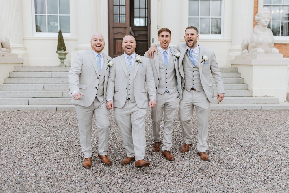 A wedding party wearing light gray suits poses together on a gravel driveway outside an elegant white building.