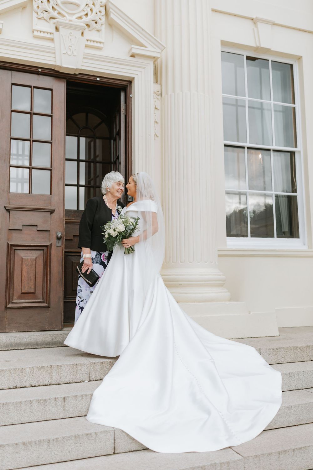 Elegant wedding couple shares romantic moments on grand stone steps of historic building with white columns and wooden doors.