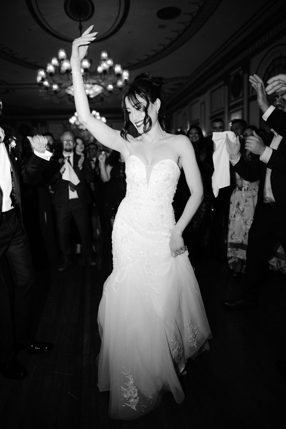 A bride in a white wedding dress dances joyfully at a dimly lit reception venue with string lights overhead.
