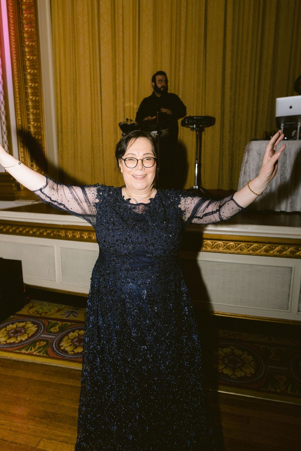 Person in black dress raises arms joyfully while dancing in an elegant room with wooden floors and ornate walls.