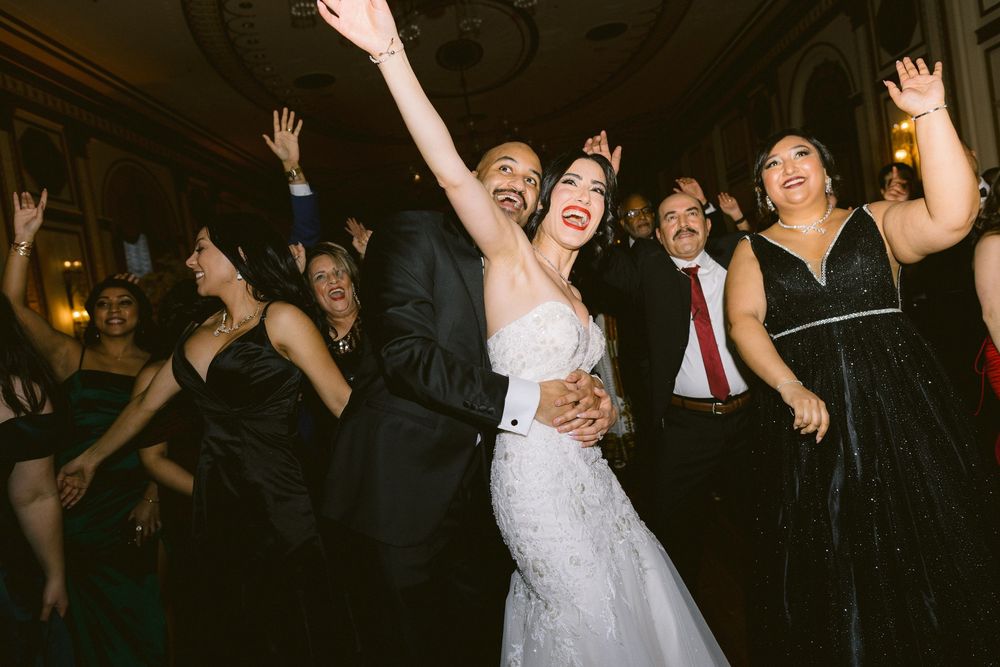 Wedding guests and bride dancing and celebrating with raised arms on a dark dance floor at a reception party.