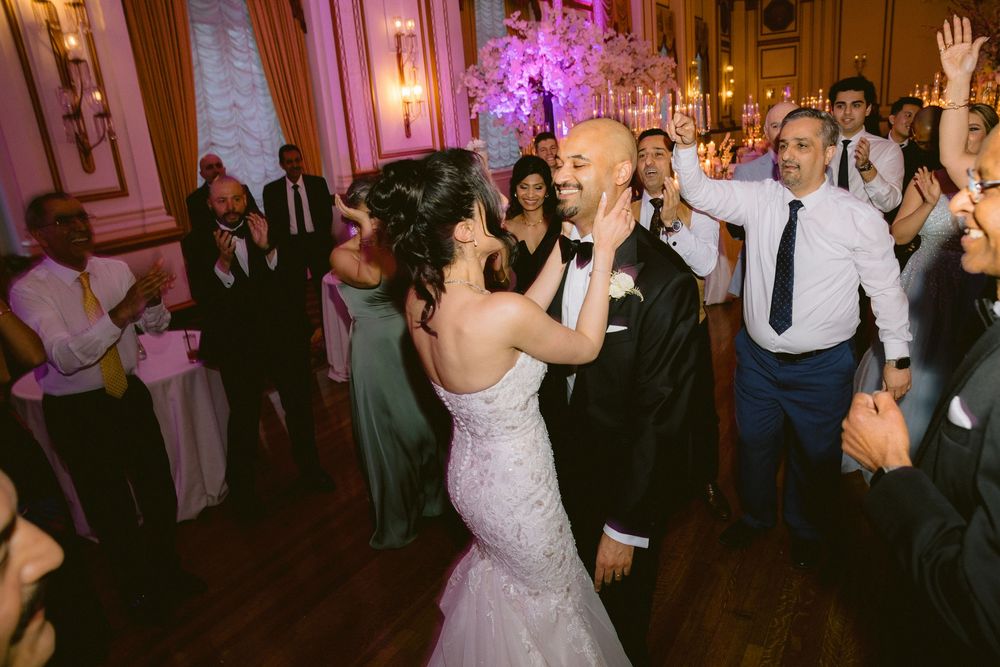 Couple dances together at an elegant wedding reception with pink lighting and wooden floors in an ornate ballroom.