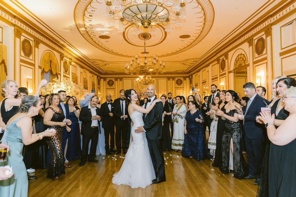Wedding guests line the sides of an elegant ballroom with ornate gold ceiling as a couple dances in the center.