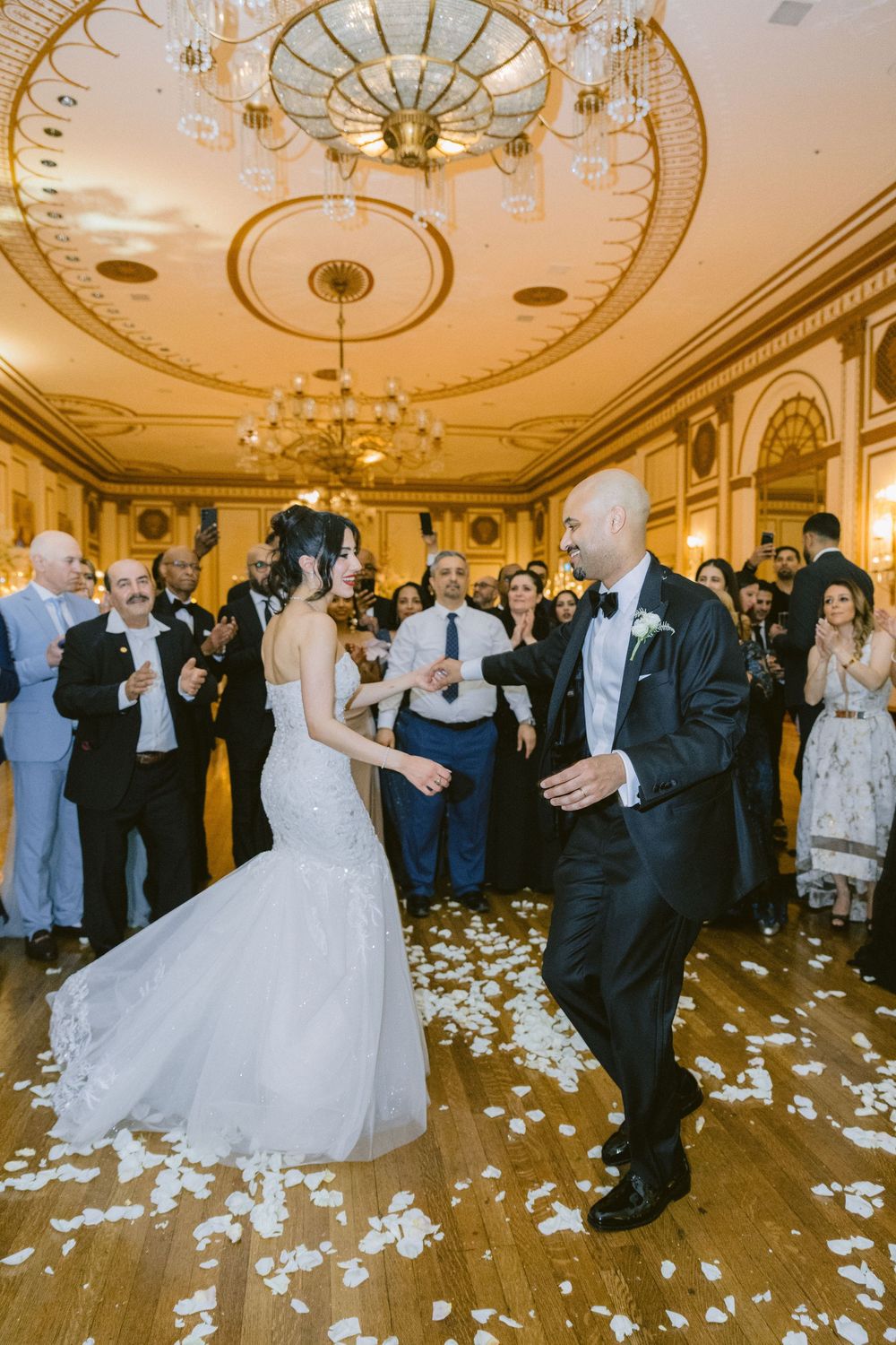 Elegant wedding reception dance in a grand ballroom with ornate ceiling and scattered rose petals on the floor.