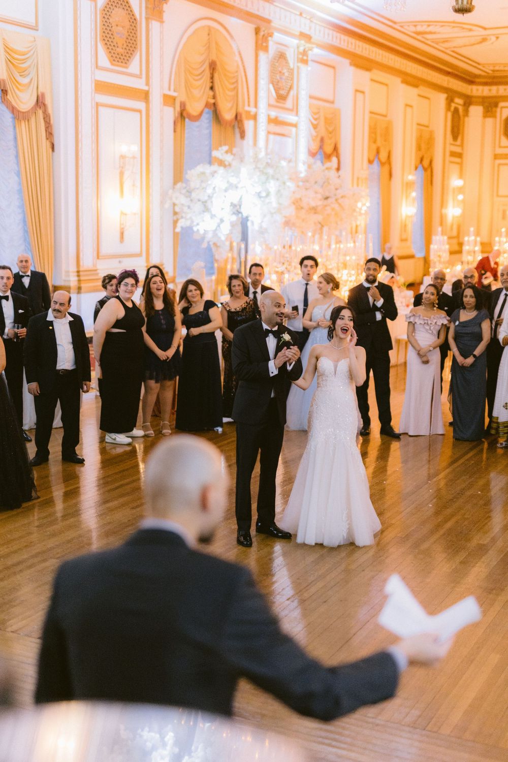 Wedding guests in formal attire gather on a grand ballroom dance floor with ornate gold decor and hardwood flooring.