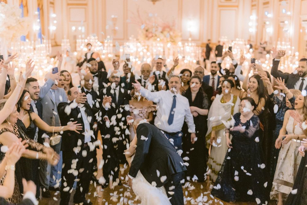 Wedding guests dance and celebrate on a ballroom dance floor while white flower petals fall from above.