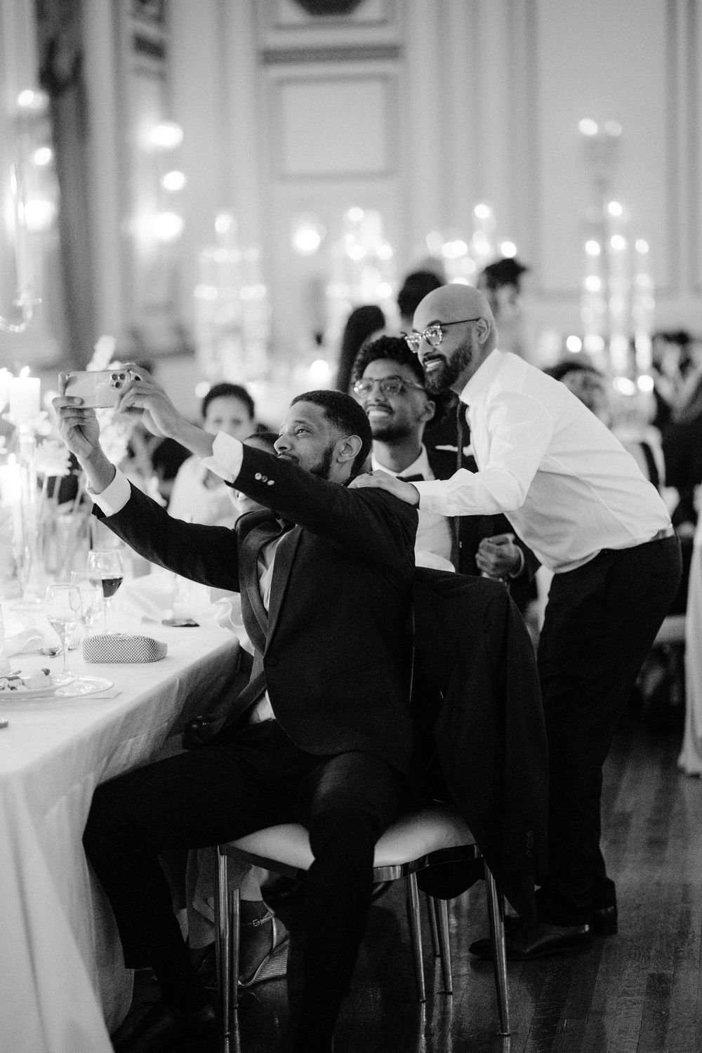 Black and white photo of guests taking selfies at an elegant dinner reception with candlelit tables.