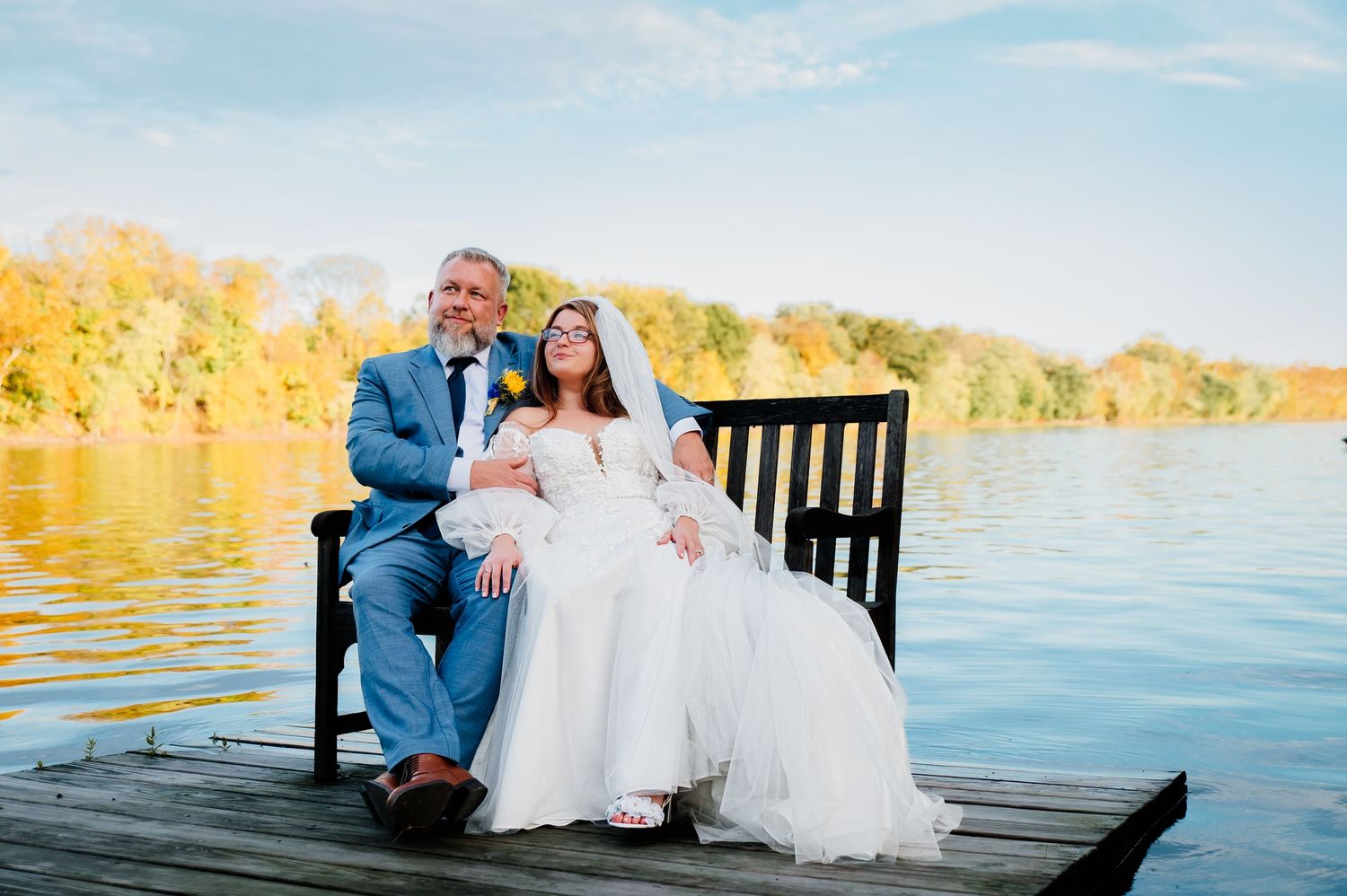 Newlywed couple sitting on wooden dock at sunset by golden lake waters.