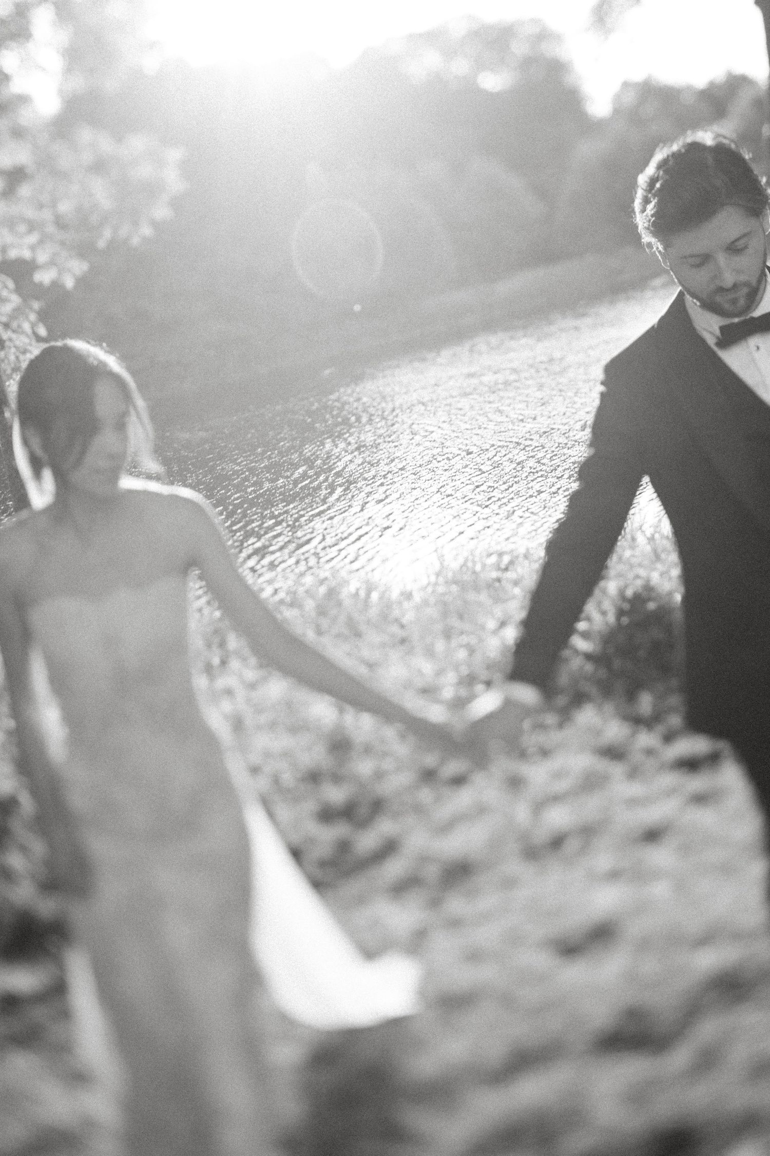 A black and white photograph shows a couple in formal attire sitting together by a reflective lake at sunset.