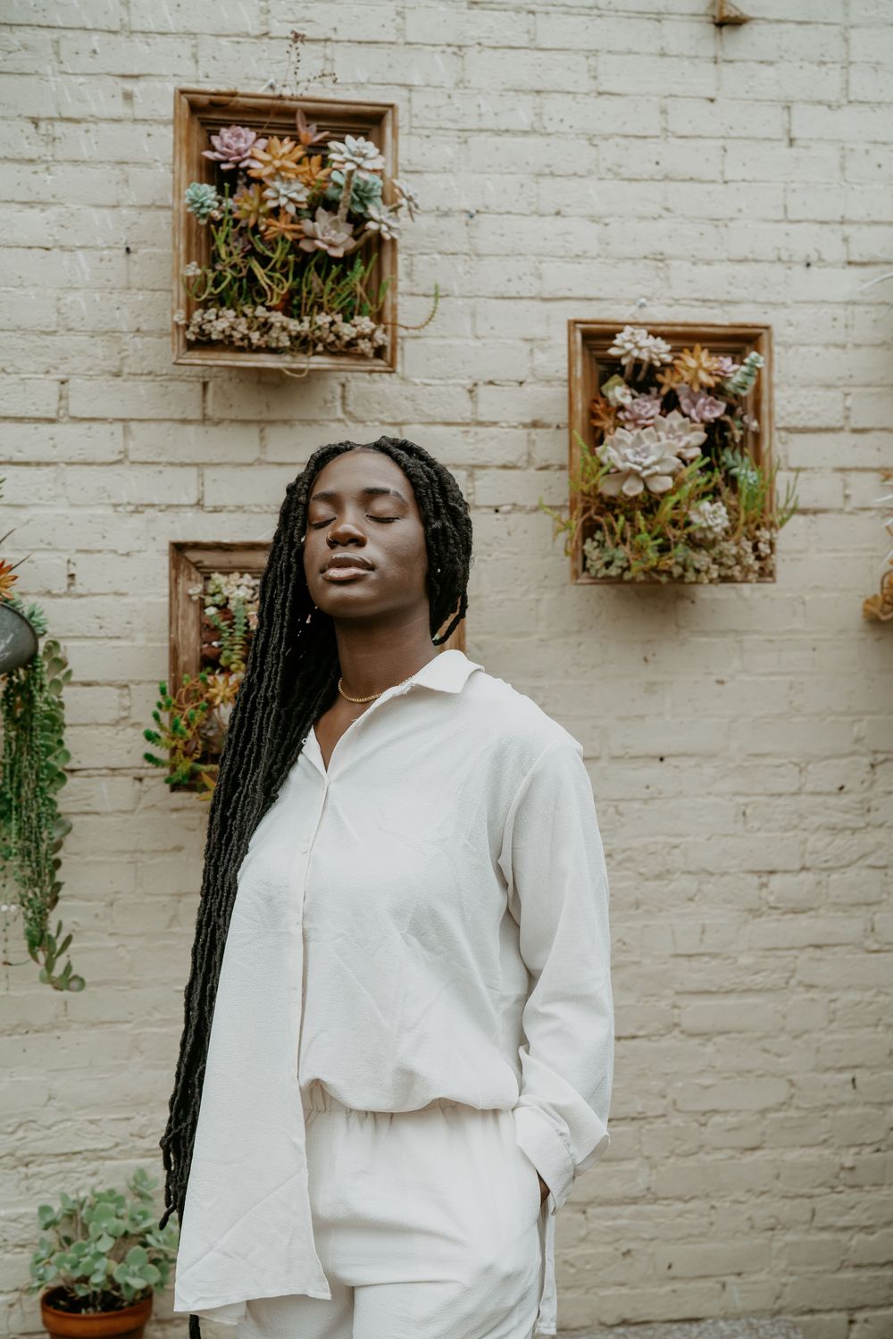 Figure in crisp white outfit poses against white brick wall with hanging flower boxes.