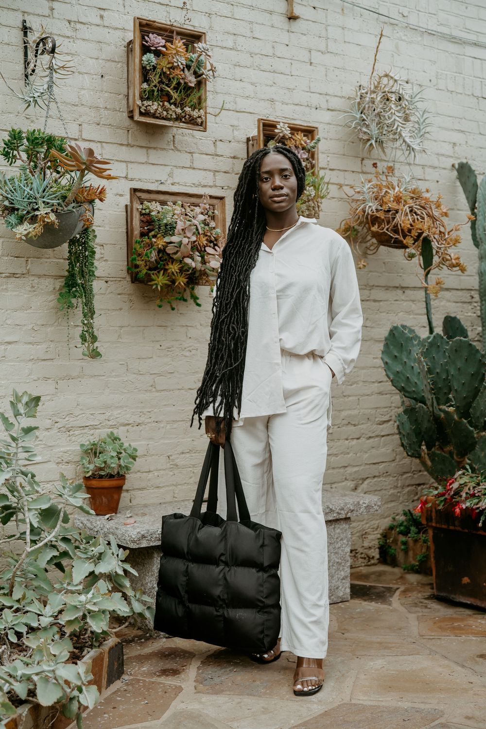 Person in all-white outfit stands near potted plants and white brick wall with black tote bag.