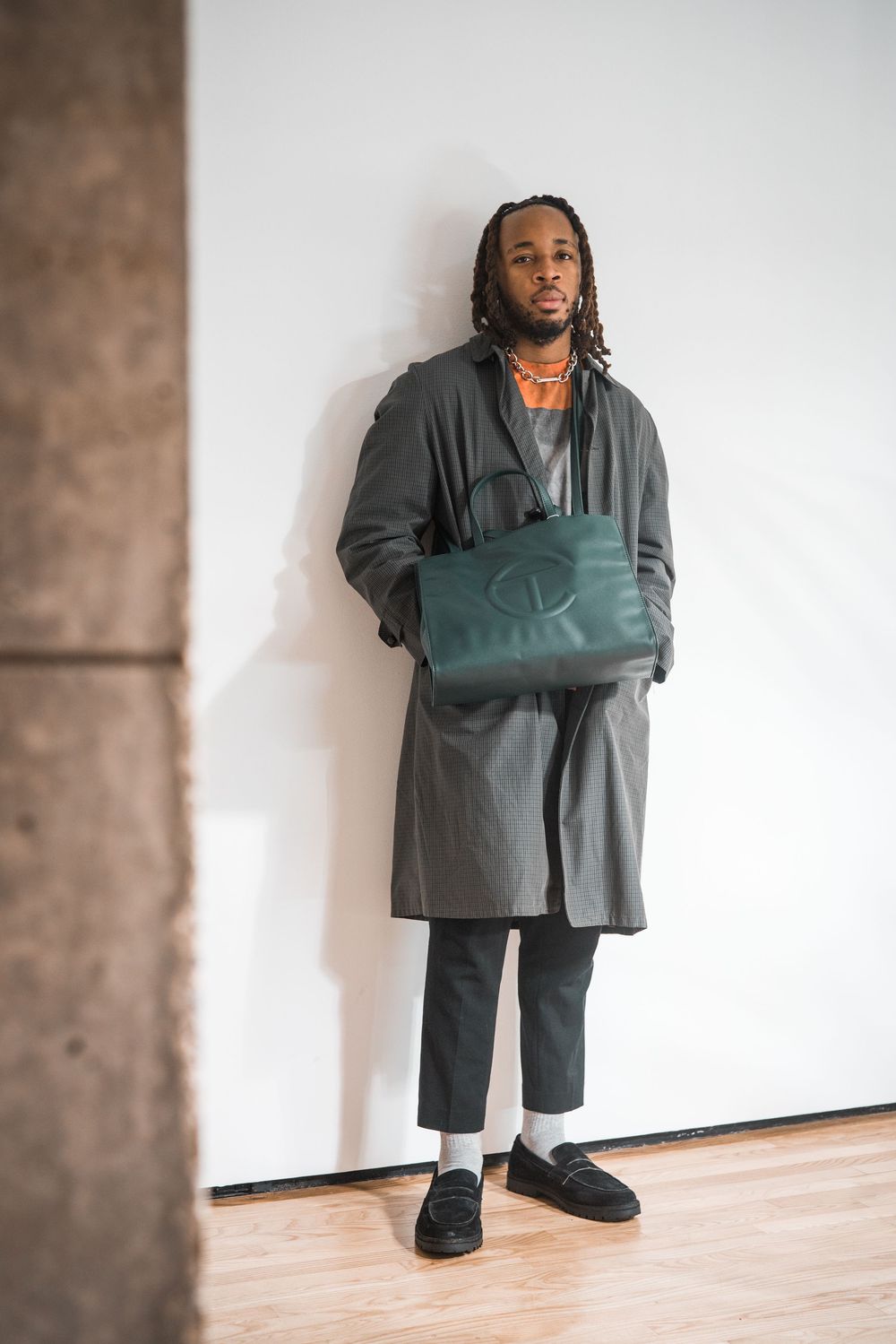 Person in dark oversized clothing poses against plain white wall in artistic setting.