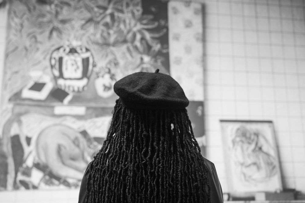 A person wearing a black beret and dreadlocks views artwork in a gallery setting.