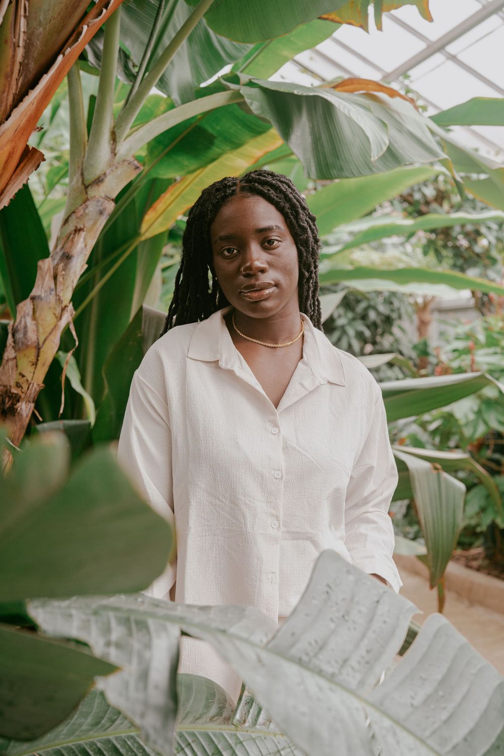 Person in white shirt poses among tropical plants and banana leaves in a greenhouse setting.