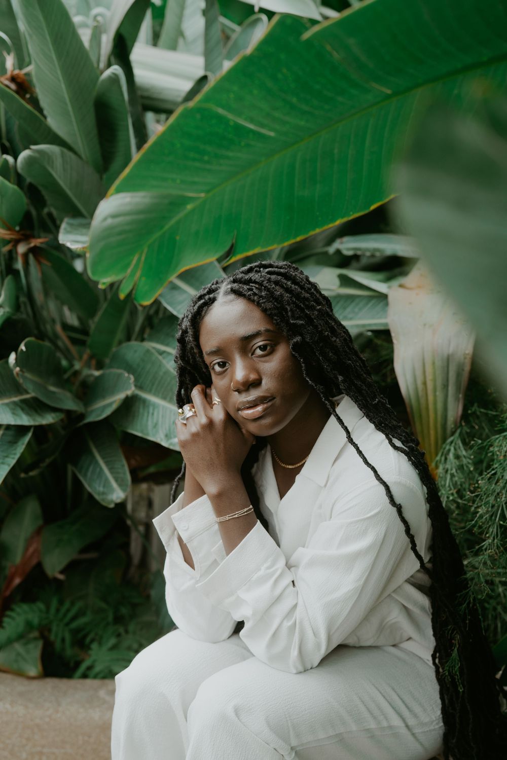 Person in white outfit seated among lush tropical plants and large green leaves.