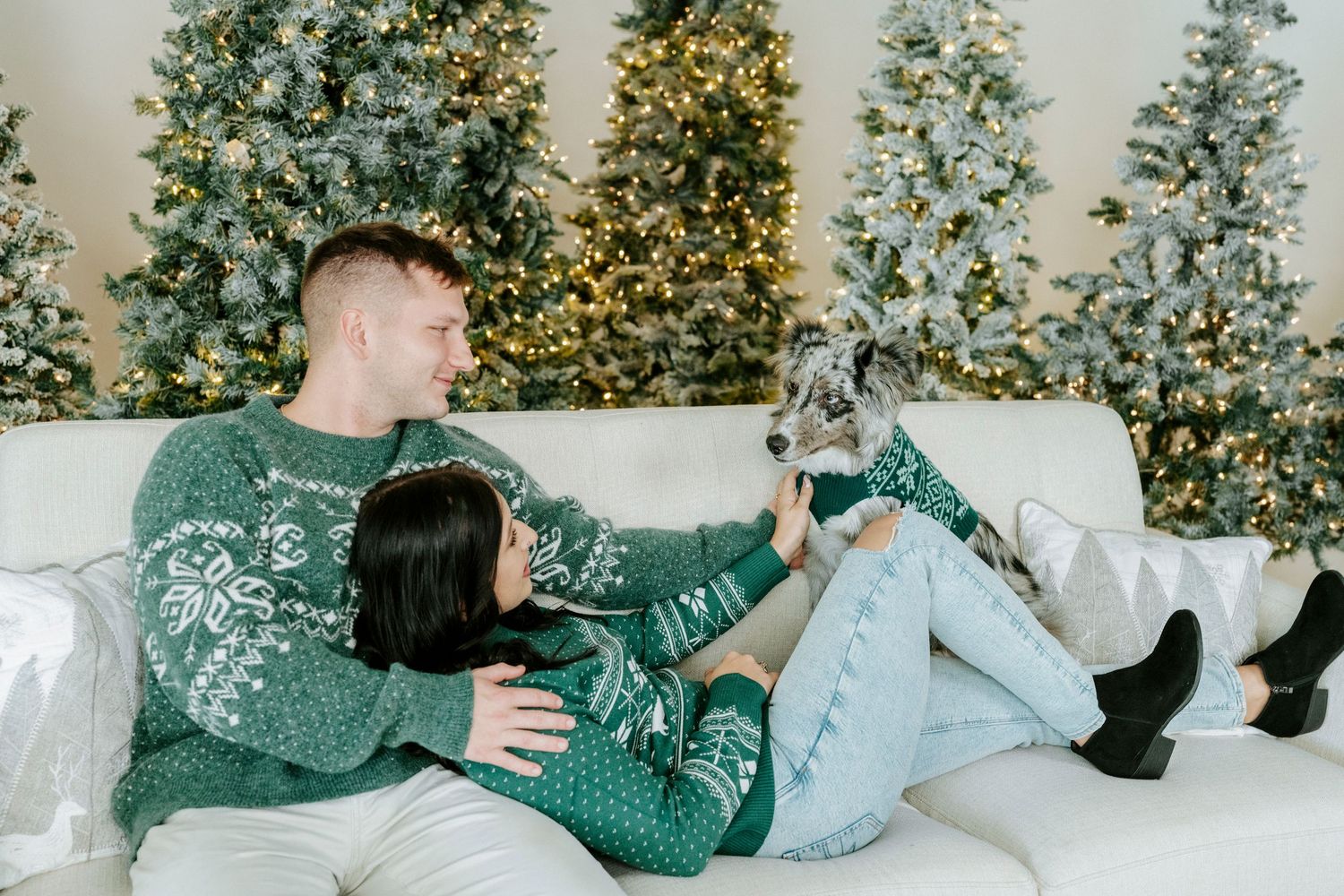 A couple in matching green holiday sweaters share playful moments on a white couch with a Christmas tree in background.